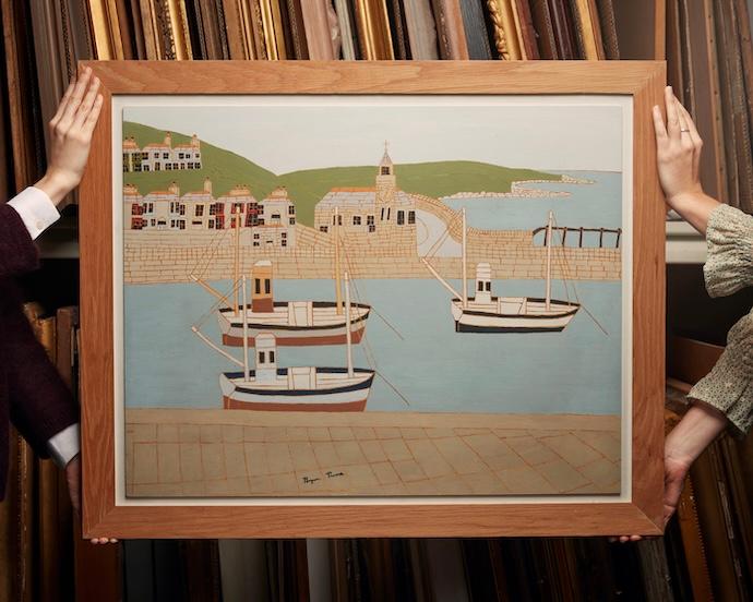 Two people hold a framed painting of boats docked at a harbour with buildings and green hills in the background, in front of shelves filled with picture frames.